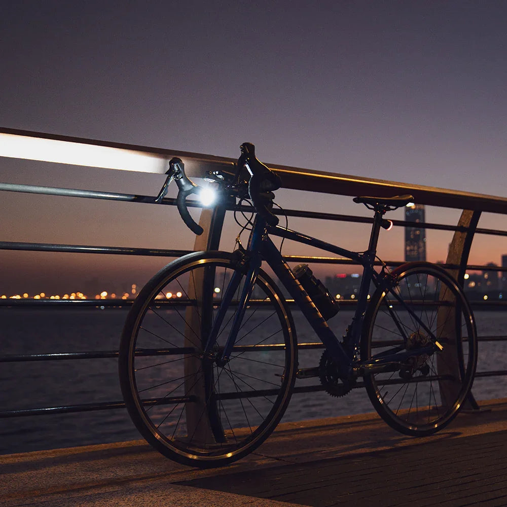 A road bike parked on a city waterfront at dusk, with its Towild CL1600 front bike light turned on and casting a powerful bright beam.