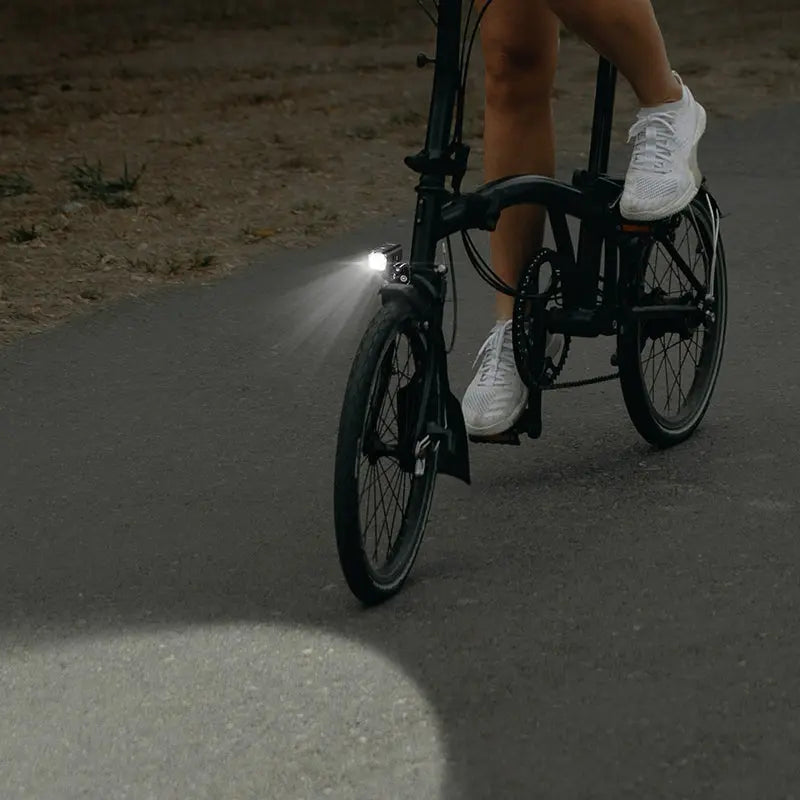 The TOWILD Urban Mini light mounted on the front of a Brompton folding bike, illuminating a path during an evening ride.